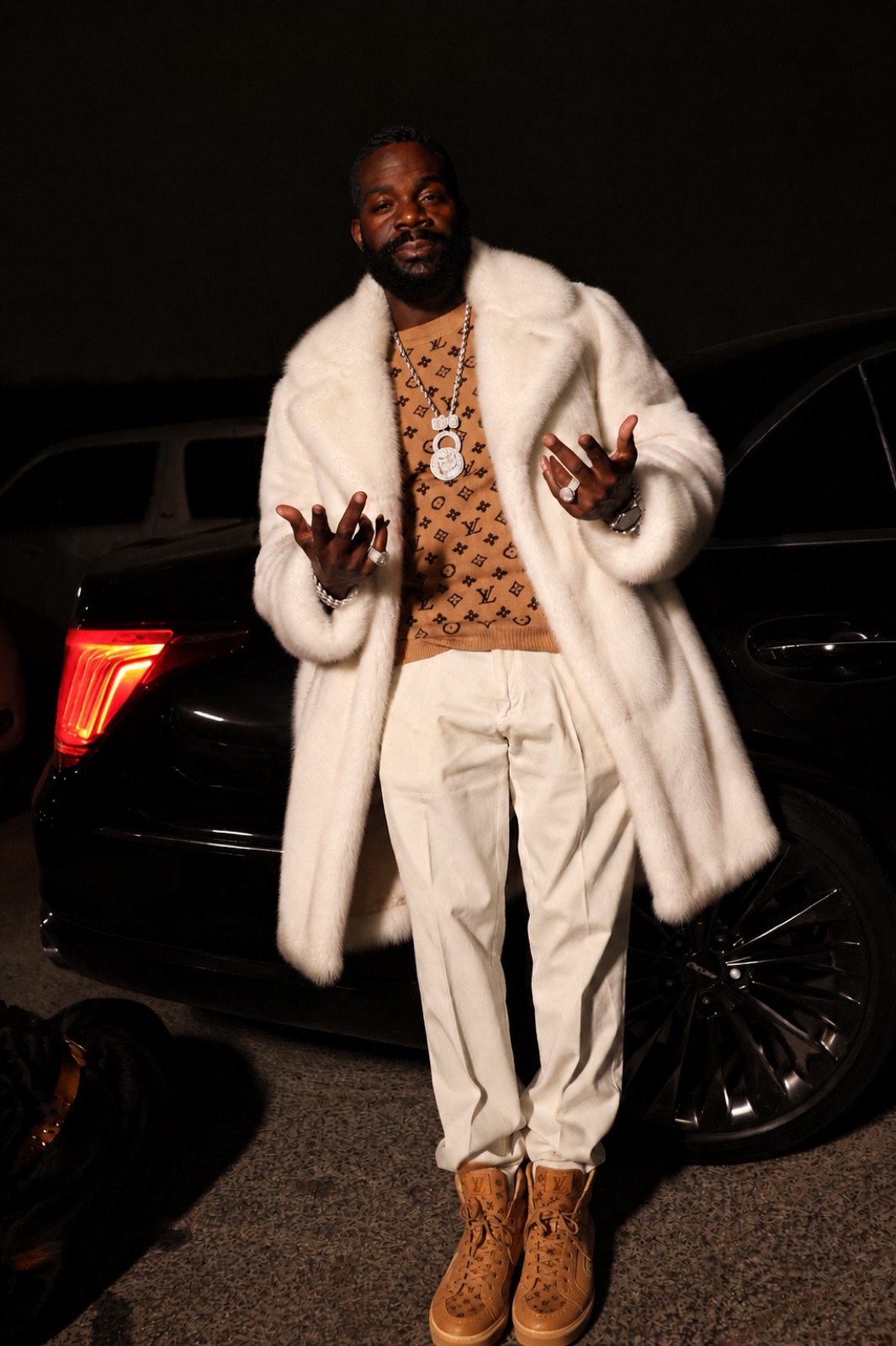 Don Chief in a white fur coat leaning on a car at night